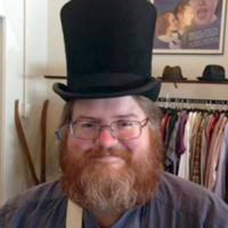 Edward is facing forward and smiling. He has red hair and a beard and wears a black top hat.