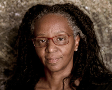 Letitia is facing forward and smiling. She has long dark hair and wears red glasses.