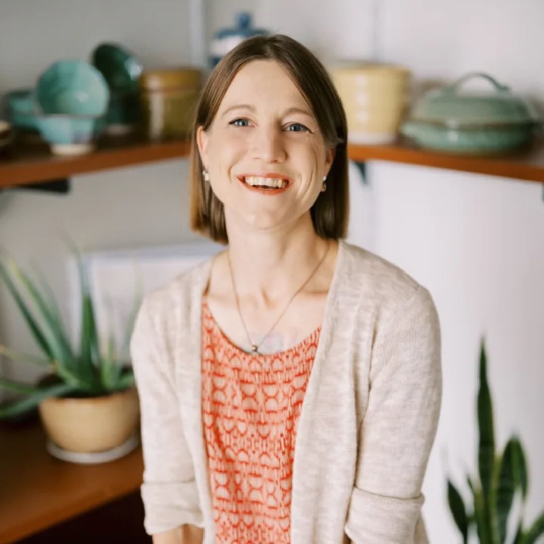 Erin is facing forward and smiling. She has shoulder-length hair, wears an orange top under a beige sweater.