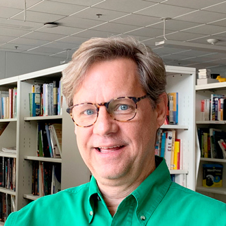 Eric Ligon smiling, brown hair, wearing glasses, bookshelves in the background