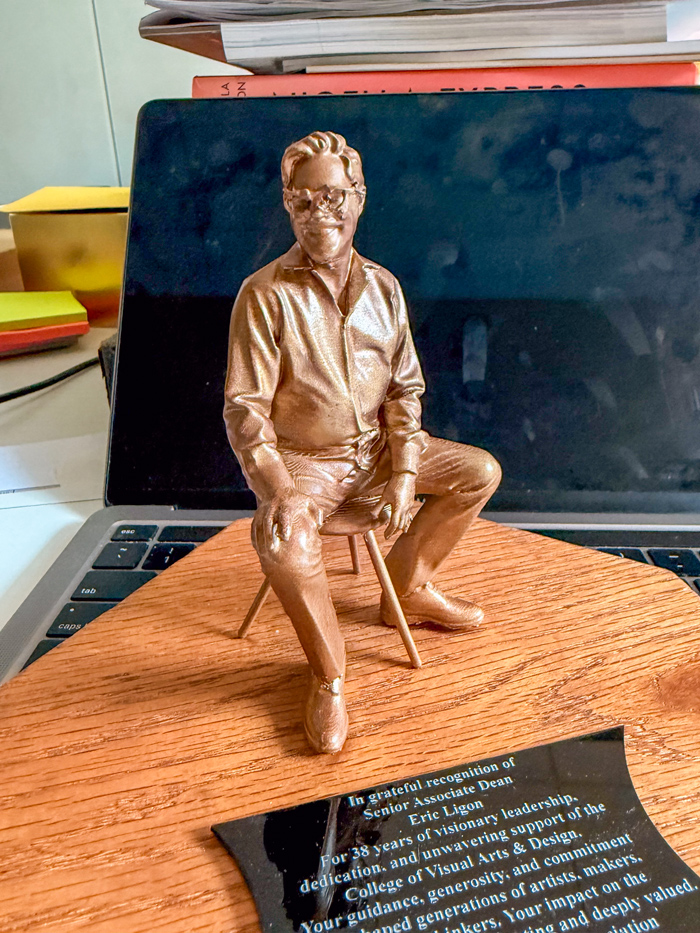 A small bronze-colored sculpture of Eric Ligon seated on a stool rests on a wooden base with an engraved plaque recognizing his 38 years of leadership and service to the UNT College of Visual Arts and Design. The detailed figurine shows Ligon smiling in glasses, with one leg crossed casually over the other.