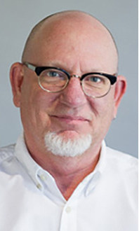 Keith is facing forward and smiling. He has white hair and wears glasses and a white shirt.