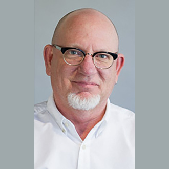 Keith is facing forward and smiling. He is bald, wears black glasses and has a white beard.