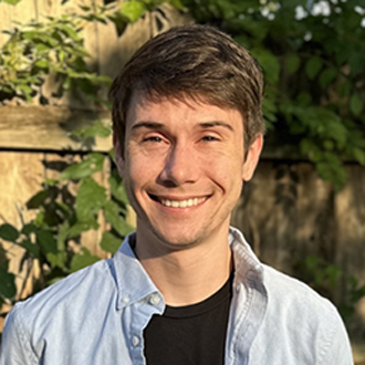 Max is facing forward and smiling. He has brown hair and wears a blue shirt.