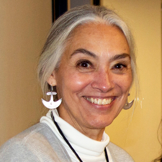 Johnnie Stark is facing forward and smiling. She has silver hair and wears a gray sweater over a white turtleneck shirt.