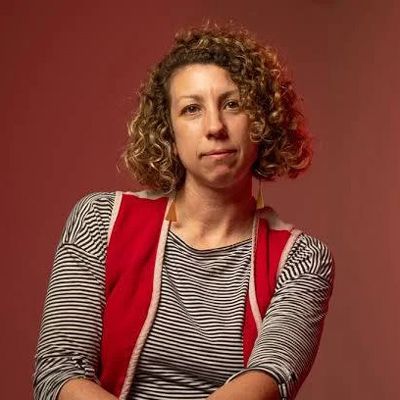 Alicia is shown from the waist up facing forward. She has curly brown hair and wears a red vest over a black-and-white top.