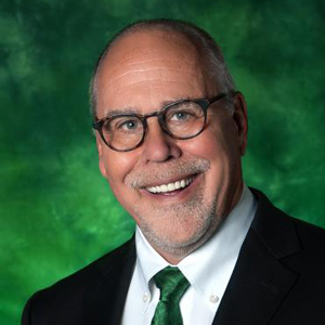 Neal Smatresk facing forward and smiling. He wears glasses and a black suit with a green tie.