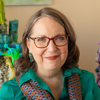 Christine is facing forward and smiling. She wears brown glasses, has brown hair and wears a green shirt with a knitted scarf.