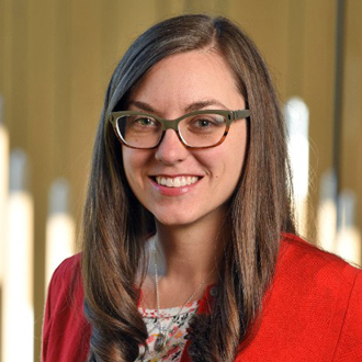 Julie facing forward, smiling, long brown hair, brown glasses, print shirt, orange jacket