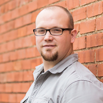 Cory smiling, shaved head, glasses, beard, khaki shirt