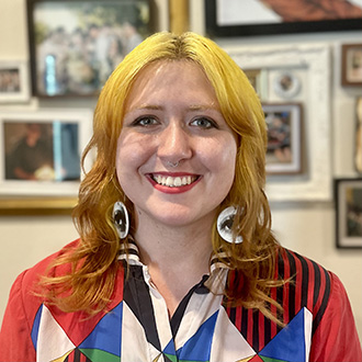 Courtney facing forward smiling, bright yellow hair, wearing a colorful shirt and large earrings with eyes on them