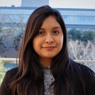 Karla facing forward smiling, long brown hair, buildings in the background, brown jacket, floral-print shirt