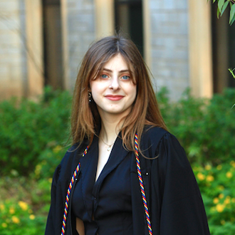 Ally facing forward smiling, long brown hair, navy blue jacket, garden in background