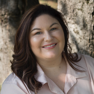 Amanda facing forward, smiling, long brown hair, pink shirt, tree in the background