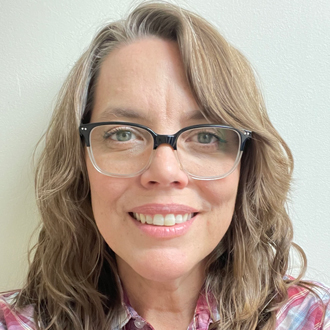 Carla with dark blonde hair and glasses, smiling and wearing a red-and-blue plaid shirt. 