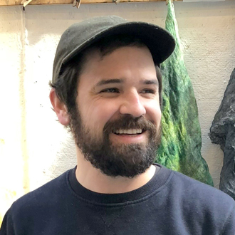 Mark Alister Raymer smiling and looking to his left, wearing a baseball cap, faded black sweatshirt