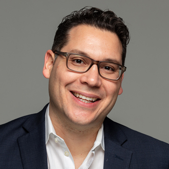 Giovanni smiling, brown short hair, glasses, white collared shirt, blue jacket