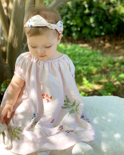 A baby sits, looking down. She is wearing a pale pink silk dress and matching hair bow that has been hand embroidered with wildflowers