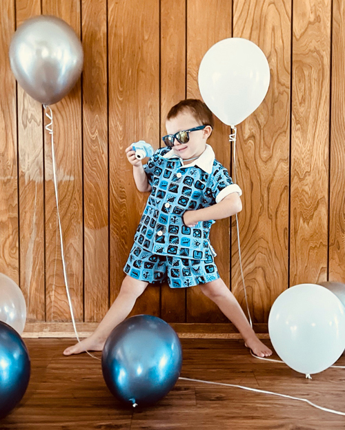 A preschooler wears a blue shirt and shorts.  He is wearing sunglasses and holding a toy camera. He is surrounded by balloons.  