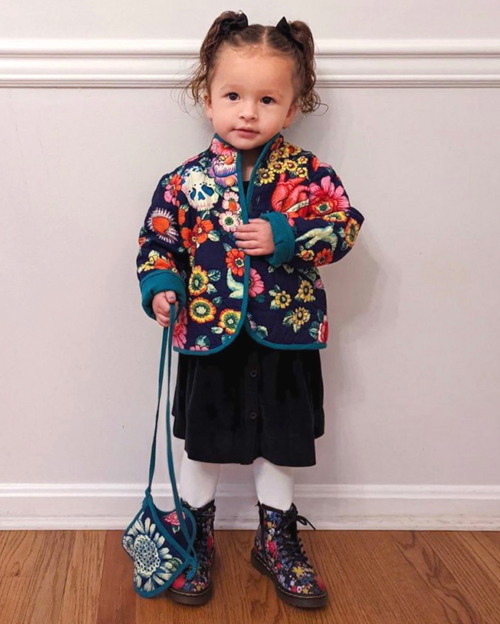 A toddler wears a colorful quilted jacket made of fabric with images inspired by the art of Frida Kahlo, with floral Doc Martens.  