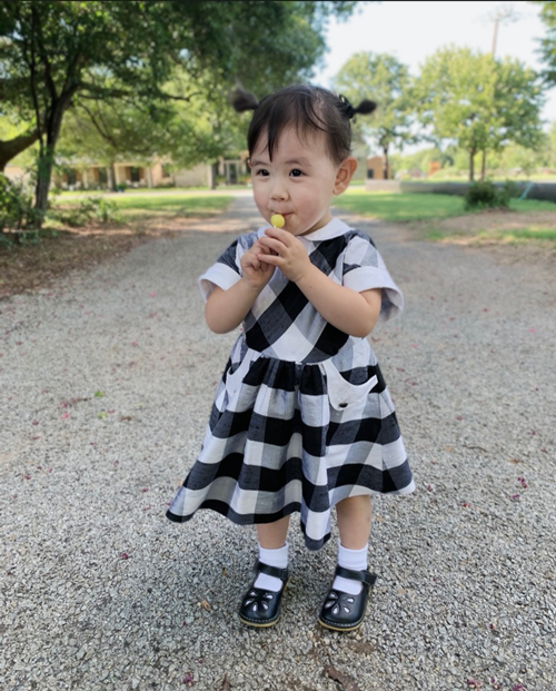 A toddler samples a yellow lollypop while wearing a black and white gingham dress made of silk.  