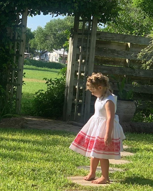 A preschooler twirls in a white and red dress. A vine-covered fence is in the background. 