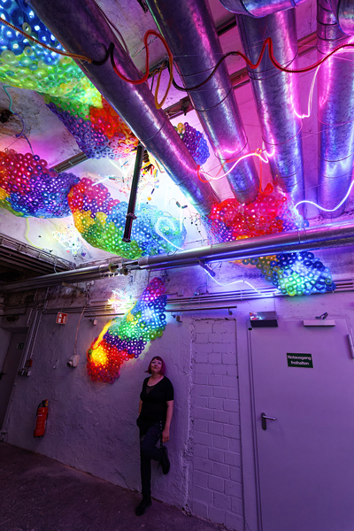 Adela standing below her light installation Chaos Exstructa. Bright lights on the walls and ceiling.