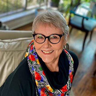 Judith Garrett Segura smiling at the camera, short gray hair, black-framed glasses, multi-colored scarf