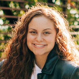 Bailey facing forward, smiling, long wavy brown hair, green jacket, white shirt.