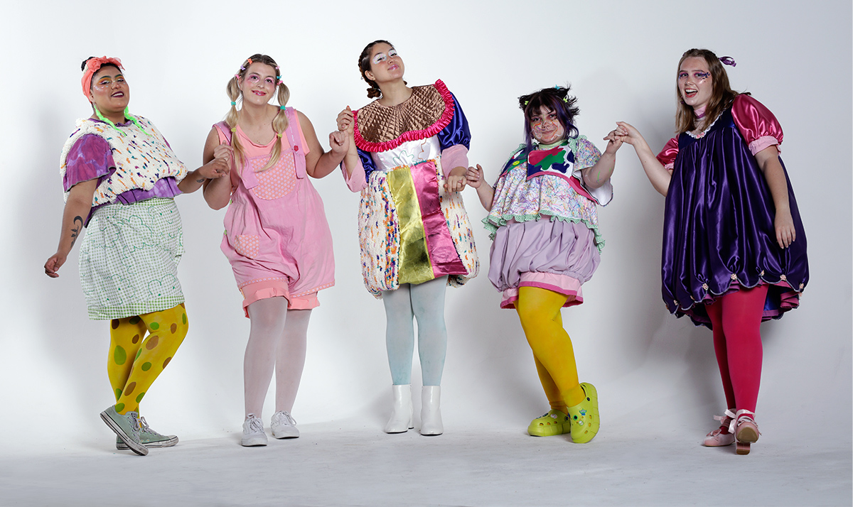 A photo of a fashion designer with her 5 models wearing her designs. On the very left is a dress, then a vest, shirt, and shorts set, then me the designer, an overalls outfit, then a dress and finally a shirt, vest, and bloomers set. Made using varying materials such as: silk, tufted burlap, cotton, muslin, spandex knit, and jersey knit. All outfits worn with colorfully dyed tights.