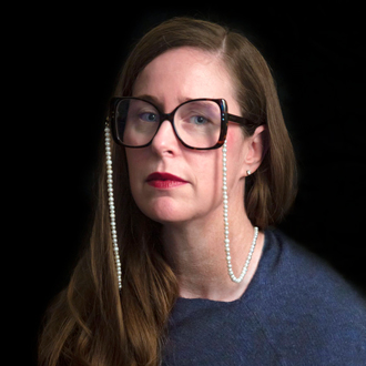 Denise Amy Baxter facing forward, wearing glasses with a pearl spectacle cord, long brown hair, blue top