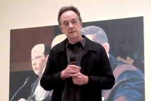 Ed Blackburn holding a microphone, older man, dark hair, wearing a brown jacket, painting in the background