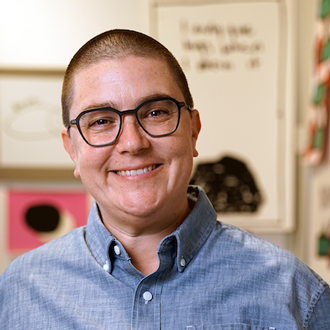 Emmy Bright smiling at the camera, closely shorn dark hair, black glasses, blue button-down shirt.