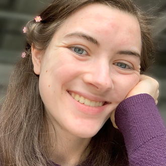 Woman smiling, brown hair, leaning on her hand, purple sweater