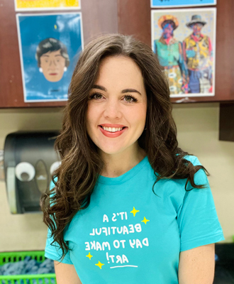 Keisha smiling at the camera, long brown hair, teal t-shirt, art in the background