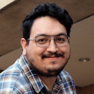 Kevin smiling at the camera, short dark hair, mustache, light beard, glasses