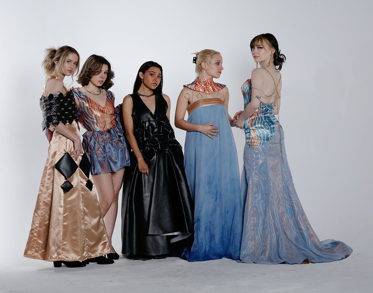 5 models against a white background, all wearing different evening wear dresses of various shades of gold, black, and blue, with gold metal adornments