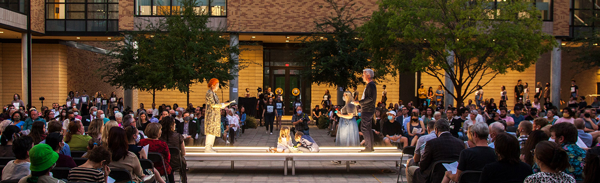 A photo of the outdoor courtyard of the Art Building, four people on a riser performing, several hundred visitors seated in attendance.