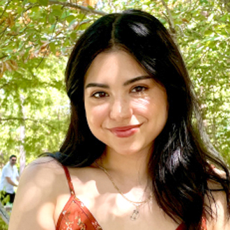 Beatriz Garza headshot. She is wearigna dress, sitting under a tree and smiling. The background is outdoors with a tree trunk visible.