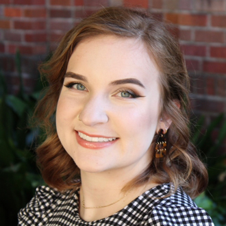 Bailey looking at the camera, brown hair, black-and-white top. Bricks in the background.