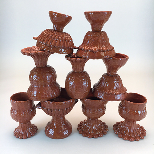Color photo of a stack of 9 wine cups with 4 on the bottom standing upright, 3 in the middle upside down, and 2 on the top upside down on a neutral white backdrop. The cups have pinched and press mold boopie glass and leafy texture with a subtle sheen on red earthenware clay. Each cup has a bowl or hourglass shape with a tall flared foot. 