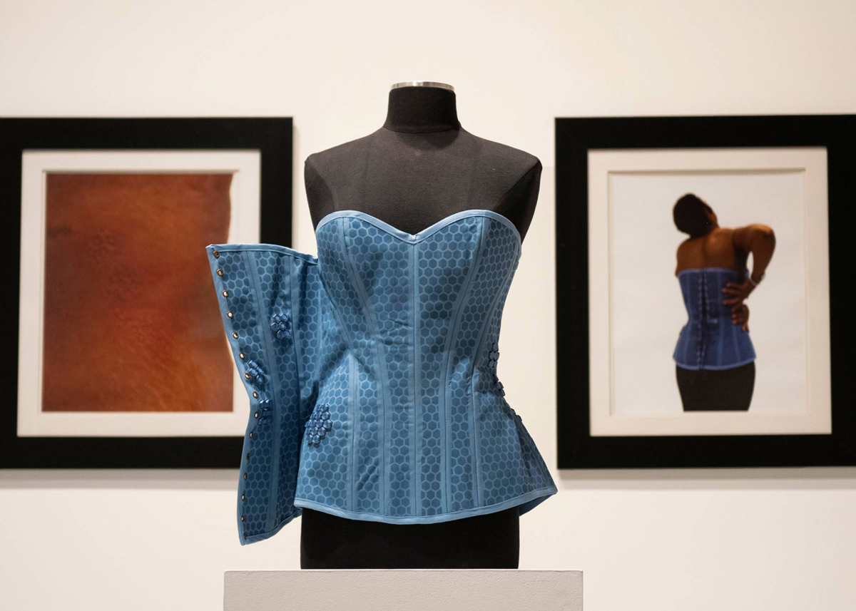 A blue corset on a black mannequin torso. Two framed pictures hang on the wall behind the torso