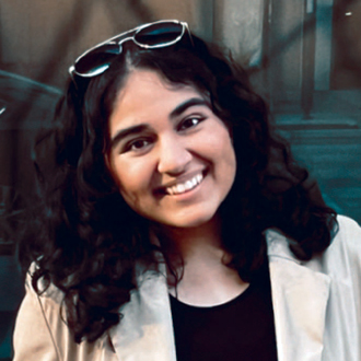 Hetvi facing forward, smiling, glasses above her forehead, black shirt, beige jacket