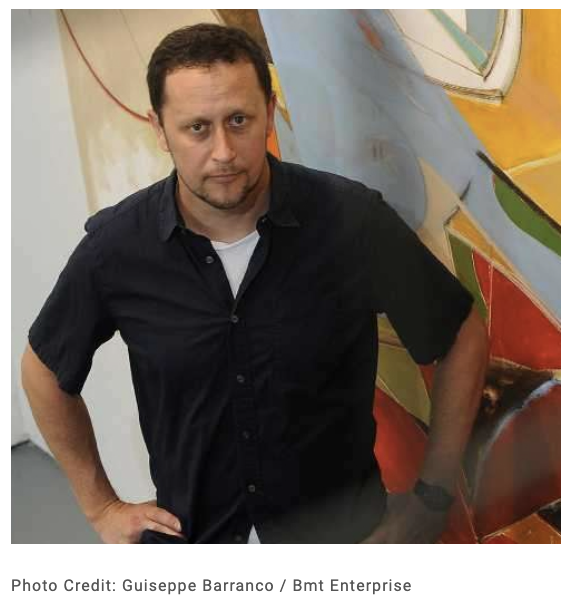 Michael Kennaugh looking at the camera, hands on hips, wearing a black shirt, painting in the background
