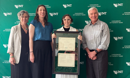 Four people facing forward smiling as one holds up a framed certificate