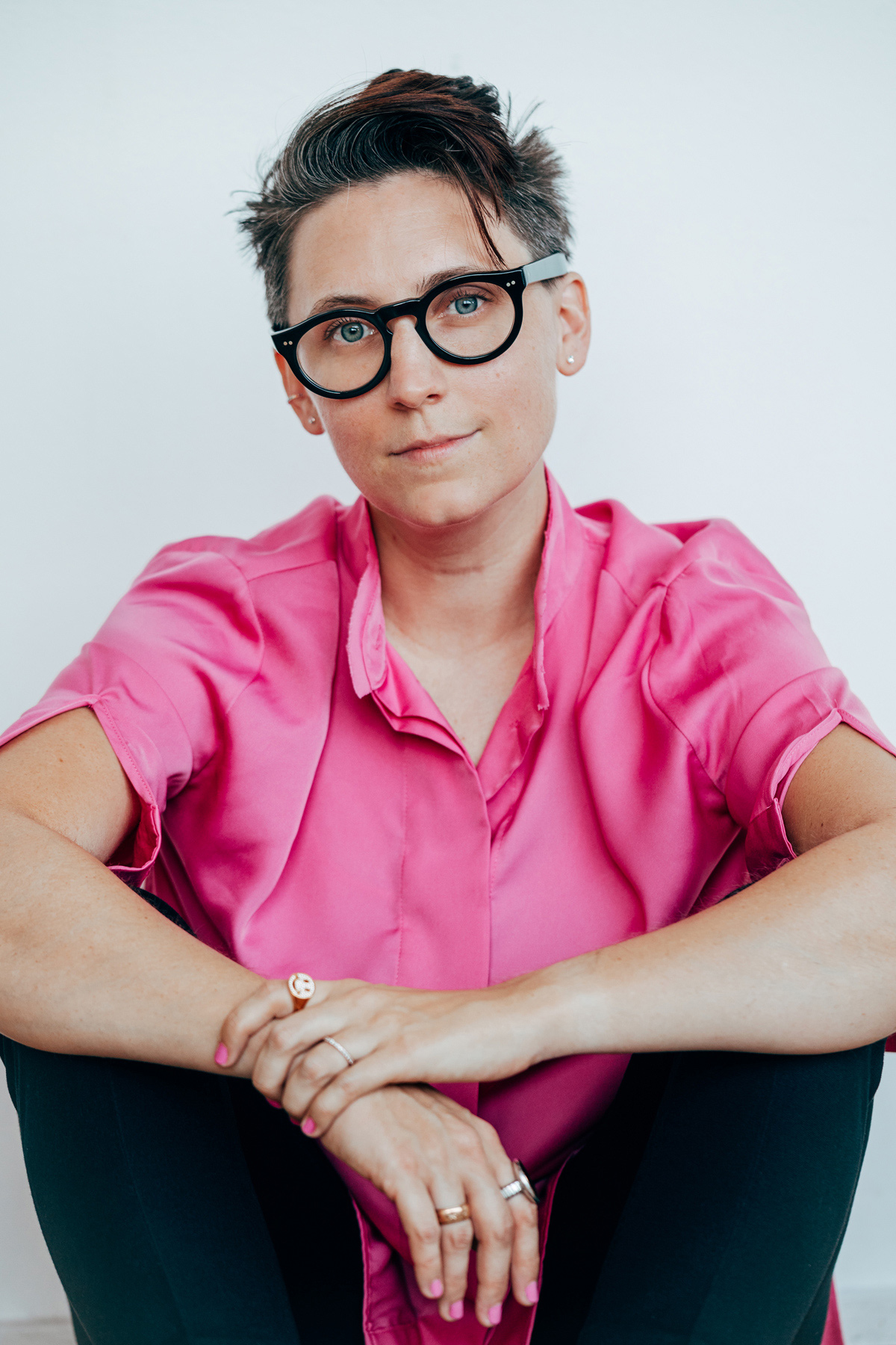 Liss LaFleur, sitting, arms crossed, wearing a bright pink shirt, black glasses