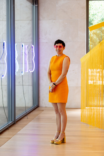Liss Lafleur, portrait, standing in the Nasher Public Gallery, smiling at the camera, wearing pink glasses, short yellow dress and gold-colored wedged sandals