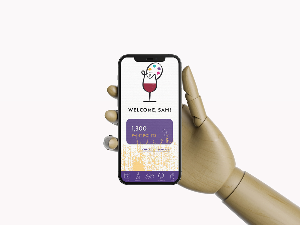 A phone is being held up with a wooden model hand. On the phone is the landing page for the Painting with a Twist rewards app. The logo sits on the top center of the landing page. Below it states: Welcome Sam! In all caps followed by the members points. 