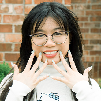 Nguyen facing forward, smiling, long dark hair, wearing glasses, holding up hands with elaborate manicure