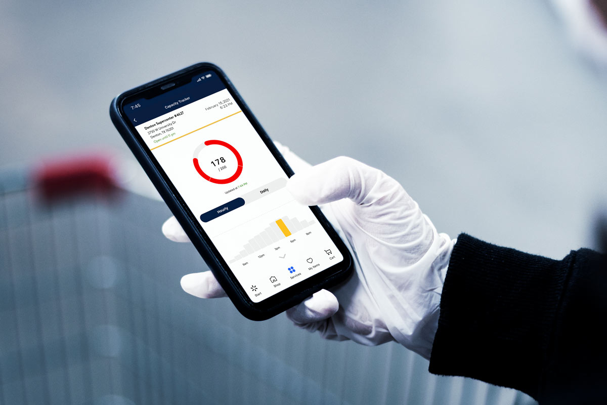 A latex gloved hand holding a mobile phone that displays the Walmart capacity tracker screen. 